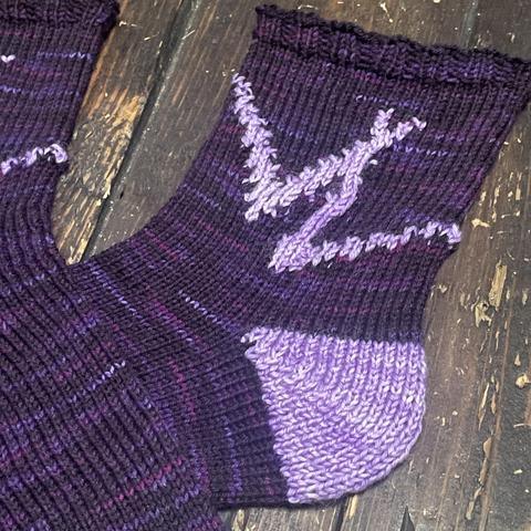 Detail of the lighter purple knitted interlocking VL pattern on the ankle of a pair of dark purple hand knit socks.