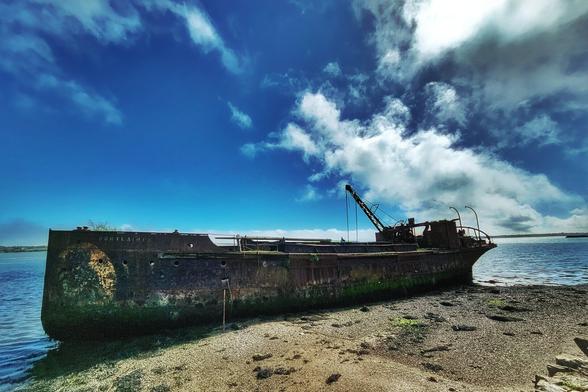 A large, rusty shipwreck rests on a rocky shore under a partly cloudy blue sky, with calm water visible in the background.