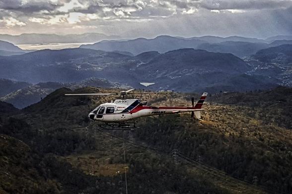 A solitary white and red helicopter hangs suspended against a backdrop of hazy, layered blue mountains under a cloudy sky, the dense forest stretching out below.