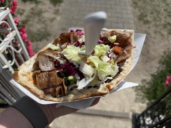 Eine Hand hält ein großes Fladenbrot, gefüllt mit gegrilltem Fleisch, frischem Salat, lila Kohl und anderem Gemüse. Im Hintergrund ist ein verschwommener Garten mit rosa Blumen zu sehen.