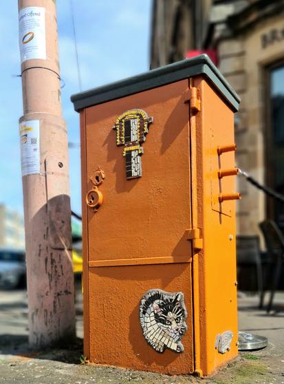 A junction box decorated with a mosaic cat chasing a mosaic mouse around its base