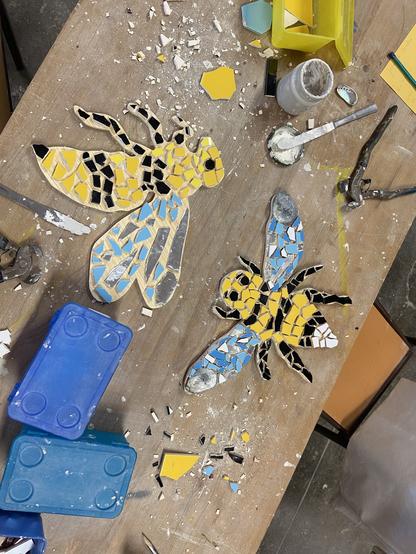 Een bij en een wesp van mozaïeksteentjes op een houten werkbank. Er omheen veel losse reststukjes en twee blauwe plastic bakjes. Er ligt ook een tang en er staat een potje met wat tegellijm.