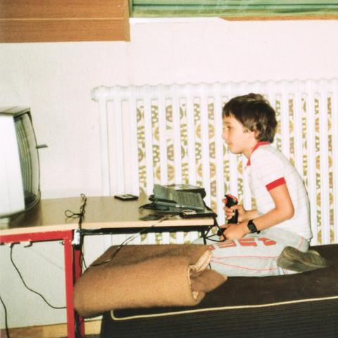 Kleines Kind hockt auf dem Sofa. Man sieht einen Tisch mit C116 und einen Ferseher. Das Kind (ich) hält einen Joystick in der Hand und starrt gebannt auf den Fernseher.