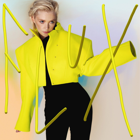 Alison Goldfrapp wearing a yellow jacket, looking through large yellow letters spelling 'Flux' in the air in front of her.