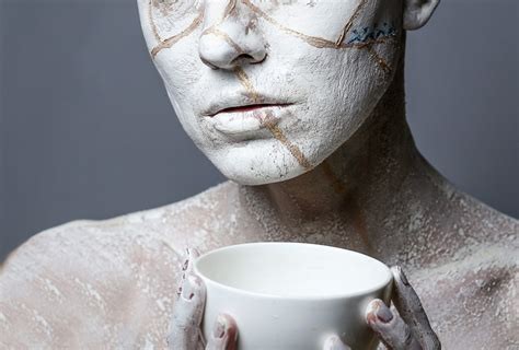 Mensch-gesicht und porzellantasse in den Händen haltend; die Tasse ist heil, das Gesicht blass geschminkt und Bruchlinien darauf gemalt, als wär das Mensch zerbrochenes porzellan.