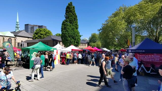 Bunte Pavillons verschiedenster Organisationen auf einem Platz, dazwischen menschen. Strahlend blauer Himmel.