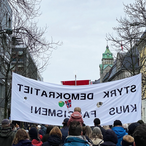 Showing a banner saying "strengthen democracy, crush fascism", but it is seen from the back (we are walking behind it. Centred in the image is the back of a child on someone’s shoulders and a series of heads.