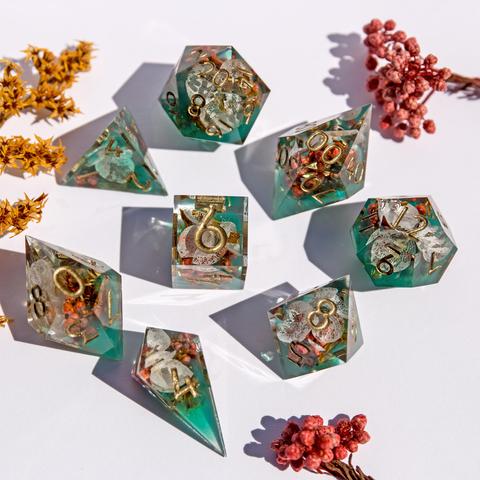 Ein Würfelset in transparentem grün mit Blüten im inneren. Das Set besteht aus einem D4-Shard, einer D4-Pyramide, D6, D8, D10, D100, D12 und D20. Das Set liegt auf weißem Untergrund mit der höchsten Seite nach oben. Die Würfel wild verteilt. Um die Würfel liegen einige rote und orangene Blüten um das Bild abzurunden.