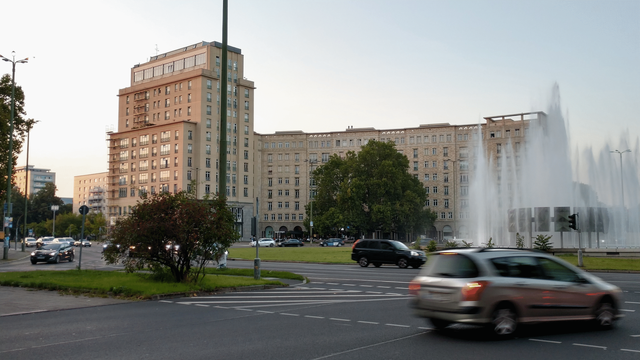 Ansicht eines westlichen Teils des Strausberger Platzes in Berlin. Im Hintergrund die Prachtbauten in Sandsteinfarben unter hellem Himmel, im Vordergrund der belebte Kreisverkehr mit fahrenden Autos. Bäume, Sträucher und ein großer Srpingbrunnen beleben den Platz.