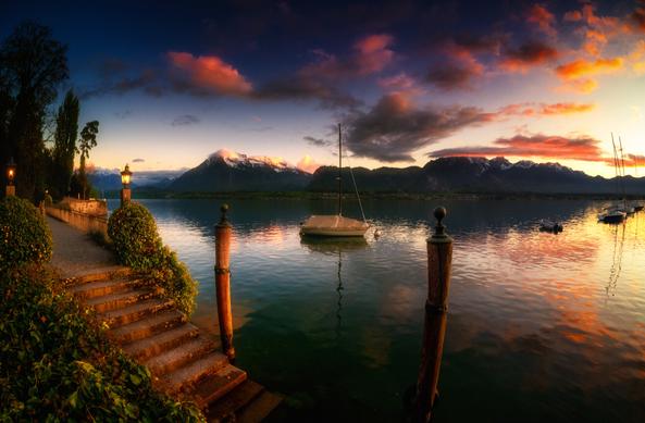 Abendstimmung am Strandweg in Hilterfingen am Thunersee. Links im Bild führt eine Treppe ins Wasser; im Hintergrund sind die Berner Alpen zu sehen, wobei der pyramidenförmige Niesen hervorsticht.