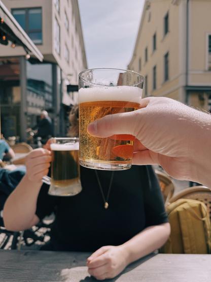 A person holds up a glass of beer, toasting with another person in an outdoor city setting under sunny skies.