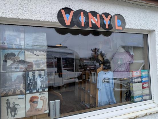 The image shows a storefront window with a sign that reads "VINYL" made from vinyl records. The window display features various items related to music and vinyl records. On the left side, there are several album covers, including images of musicians and album artwork. The center of the window displays a mannequin wearing a light blue t-shirt with the "oasis" logo, and a purple t-shirt with the word "BURNLEY" on it. To the right, there are stacks of CDs and vinyl records, along with a black cap. The background shows a reflection of a white camper van and a tree. The store's exterior wall is white with a textured finish