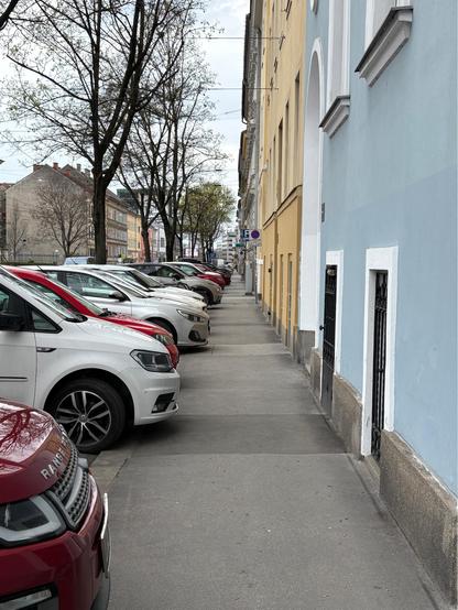 Ein Gehsteig mit mehreren parkenden Autos auf der linken Seite, die über den Gehsteig ragen, eine Mehrparteinhäuserzeile auf der rechten Seite
