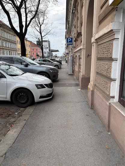 Ein Gehsteig mit parkenden Autos auf der linken Seite, die über den Gehsteig ragen und wenig Platz für Fußgänger:innen bietet