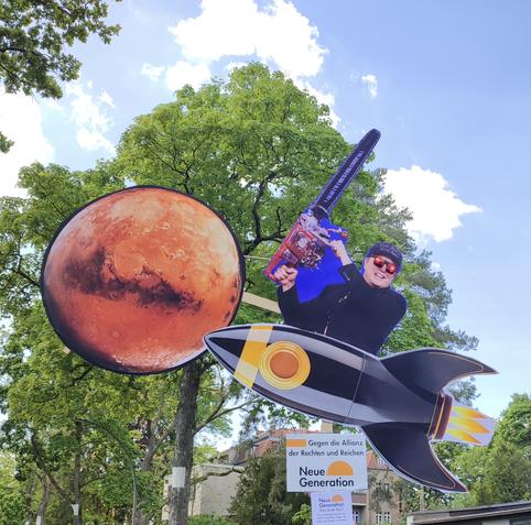 Demoschild in Form einer Rakete, die zum Mars fliegt. In der Rakete sitzt Elon Musk, er hat eine Kettensäge in der Hand und rot leuchtende Augen