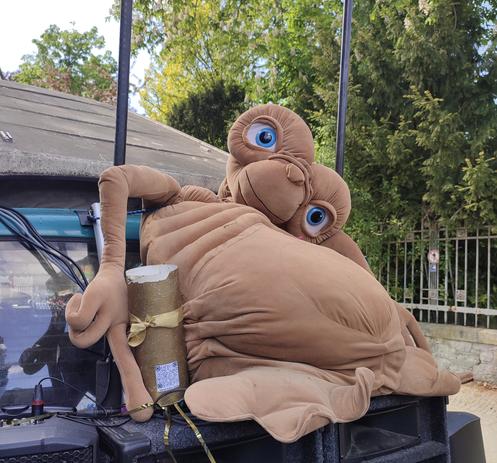 Ein sehr zerknautschter riesiger E.T. aus Stoff ist auf der Motorhaube eines Autos festgemacht