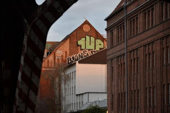 Graffiti 1UP in grün mit schwarzen Außenlinien auf der Stirnseite eines Backsteingebäudes, das etwas höher ist, als das davor stehende, weiße Gebäude. Unter 1UP noch ein schwarzes Graffiti mit weißen Außenlinien. Dort ist BIONE zu erkennen.