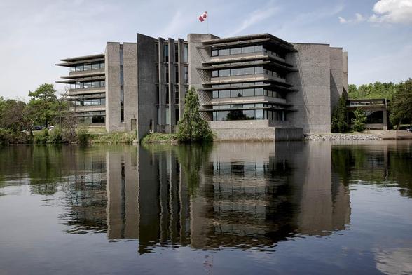 A concrete, modernist building stands by the water’s edge with its reflection visible in the river. A Canadian flag is on the roof, and trees surround the structure under a partly cloudy sky.