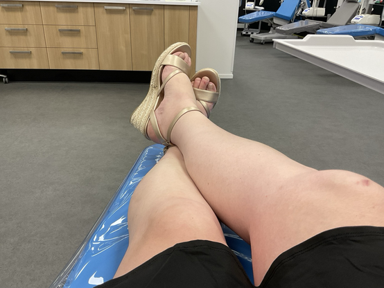 Emma zit ontspannen met haar benen gekruist tijdens het doneren van plasma bij de bloedbank. Ze draagt comfortabele sandalen met hakken en rust uit op een behandelstoel.