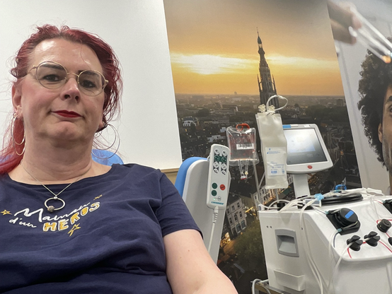 Emma, met rood haar en bril, glimlacht ontspannen tijdens haar plasmadonatie bij de bloedbank. Achter haar zijn medische apparaten te zien die gebruikt worden voor het afnemen van plasma. Ze draagt een donkerblauw shirt met een positieve boodschap.