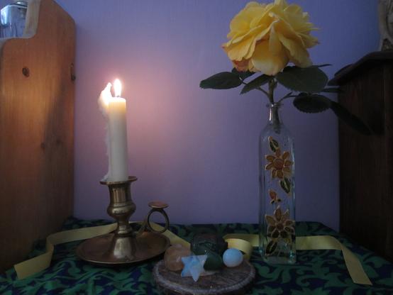 My Bealtaine altar. A White candle in a brass candle holder burns on the left. A yellow rose (Athur Bell) sits in a thin glass bottle decorated with a floral design on the right. Some crystals sit on a circular grey stone object in the middle. All sits on a blue and green meterial cloth and there is a yellow ribbon draped amongst.