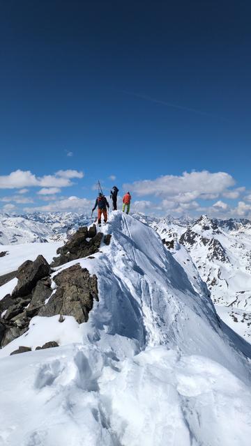 The summit ridge of the Scalettahorn. There are three people on the summit, one of them walks in my direction. The ridge is snow covered and rather narrow. A little bit below the very top some dark grey rocks are visible.