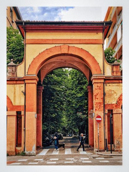 A very tall yellow, orange and brown arched/arched walkway. There are two people under the arch. One with brown hair walking towards the left, holding a blue bag and a dog and the other with grey hair walking towards the right. Through the portico you can see a street lined with big leafy green horse chestnut trees.