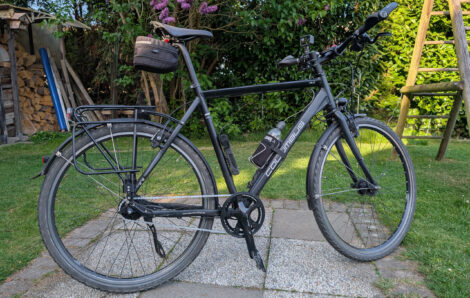Ein schearzes Fahrrad steht auf einer Waschbetonplattenfläche in einer umgebenden, grünen Rasenfläche. Im Hintergrund sieht man einen blühenden Fliederbusch.