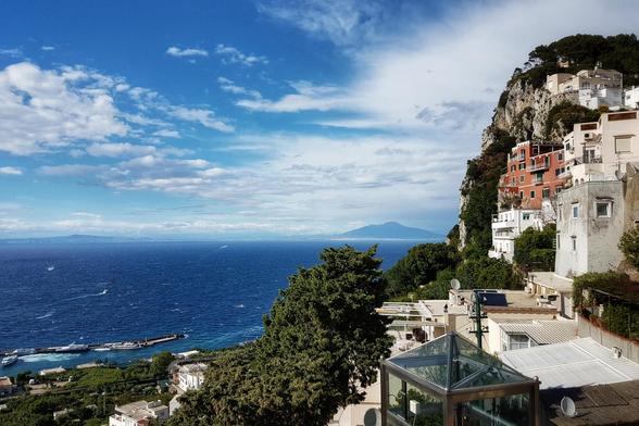 A high-angle shot shows a vibrant blue sea meeting a rocky cliff face dotted with white and terracotta buildings under a partly cloudy sky.