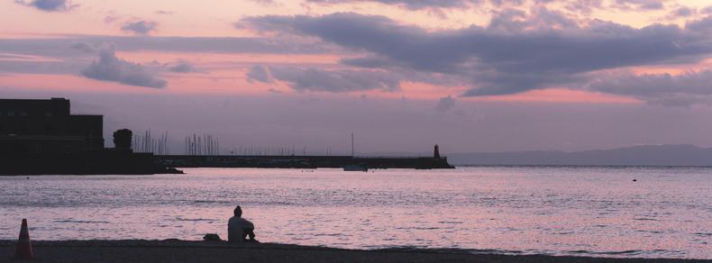 The image depicts a tranquil beach scene at sunset or dusk. There is a person sitting by the shoreline, looking out at the water. The sky is painted in shades of purple and pink with scattered clouds. In the distance, structures like a breakwater or pier are visible, along with some silhouetted buildings. A traffic cone is on the sand near the water.