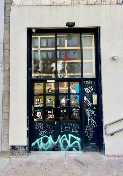 Photo of a high rectangular building entrance consisting of double doors with tall transom windows, framed in black and covered in graffiti. The doors have a grid of glass panels on their upper halves. The windows above are almost as tall as the doors themselves, and reflect the rows of windows of the building across the street (behind camera). The central window displays the number “23” in large red numerals. Text graffiti covers the lower doors and the narrow side panel to the right, as well as part of the upper windows; the text is mostly in white or black with a scattering of red and other colors, though the most prominent graffiti is “Tomas” in large turquoise letters at the bottom of the doors. The doors are set in a light-colored building wall with a vertical strip of brown panels at left frame and a horizontal strip of decorative trim at top frame. There’s also a handrail attached to the building wall at lower right. The overall effect is of grid patterns echoing and complementing each other.