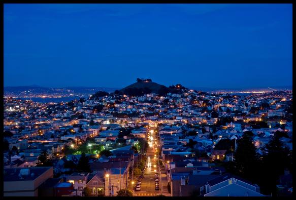 A cityscape view taken just after sunset, of 30th Street and Bernal Hill, south of San Francisco's downtown. There are some short light trails from cars on 30th Street. A bit of San Francisco Bay shows in the distance... and a little bit of Oakland as well. 

The whole scene is royal blue, with amber street lights hands a little bit of green lighting up the Upper Noe Valley playground on the left.