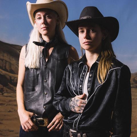 Two women stand against a canyon backdrop. On the right, a white woman with long leached hair, a cowboy hat, a leather vest, and black kerchief tied around her neck. On the left, a shorter woman with a  black cowboy hat, black western shirt, and tattooed hands.