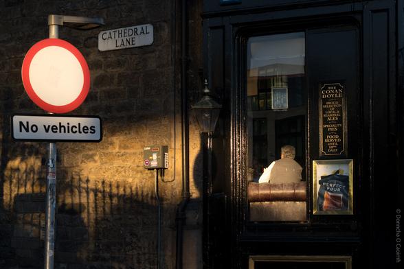 A warmly lit street scene in Edinburgh, Scotland, showing a "No vehicles" road sign and a street name plaque reading "CATHEDRAL LANE" on a stone wall. To the right, a man sits inside a pub, visible through a window with signage that reads: "THE CONAN DOYLE PROUD SELECTION LOCAL & SEASONAL ALES SPECIALITY PIES FOOD SERVED UNTIL 10PM DAILY." Below this is a poster with the text "PERFECT POUR." The evening sunlight casts long shadows across the scene.
