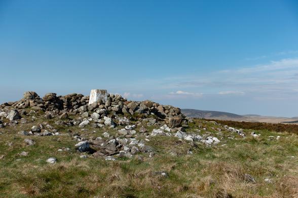 Colour photograph of a faded white trig point in a jumble of stones. There is clear blue sky overhead.