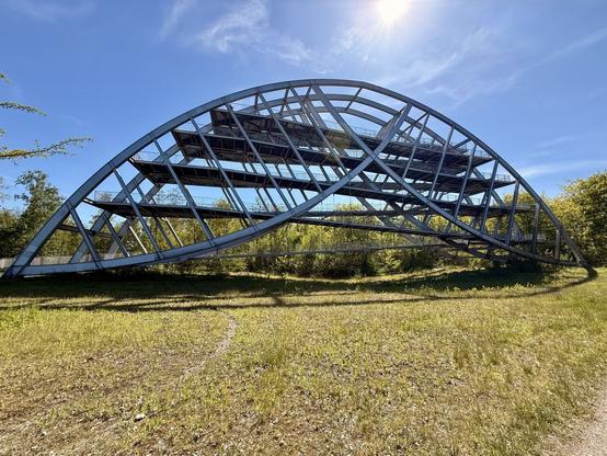 Eine große, geschwungene Metallstruktur mit mehreren Ebenen und offenen Räumen, die auf einer von Bäumen umgebenen Wiese unter blauem Himmel liegt.