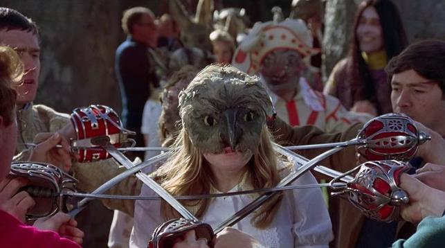 A pagan ritual. Six men with swords have them crossed so that there is a single opening in the middle. People move underneath, then stand up so that their head is inside the circle. Shown here is a young woman in an owl mask with her head in the circle, as a parade waits behind them
