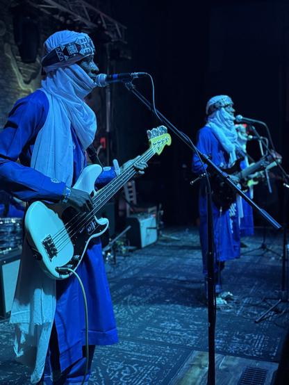 Etran del Aïr in traditional attire perform on stage. One plays an electric bass guitar and sings into a microphone, while the other stands nearby with a similar instrument. The setting is illuminated with stage lights, creating a vibrant atmosphere.