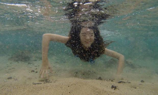 Underwater, a girl holds out her arms to skim along the surface of the sand.