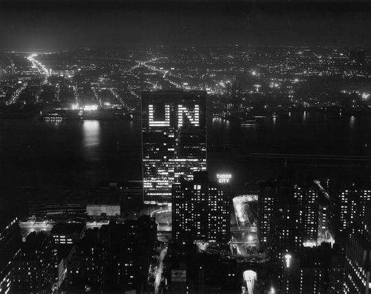 Foto van het Hoofdkwartier van de Verenigde Naties aan de East River gezien vanuit de lucht met op de achtergrond de verlichte stad New York. Door verlichte vensters van het 39 verdiepingen tellende gebouw zijn de letters UN zichtbaar.