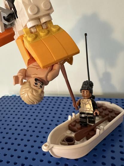 Lego MerStede, looking terrified, is hanging from the beak of a large pelican flying over the ocean. Lego Ed is below him in a rowboat, looking concerned and brandishing an oar at the bird.