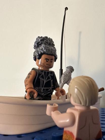 Lego Ed leaning over the edge of his rowboat to talk to Lego MerStede, who is popping out of the ocean and facing away from the camera. A fish hangs from a fishing rod in the background.