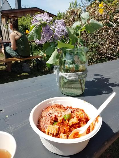 Auf einem Bierzelttisch stehen eine Pappschale mit Spirelli-Nudeln und Bolognese, davor ein Pappbecher mit einer gelben Flüssigkeit, dahinter ein Weckglas mit ein paar Zweigen Flieder. Am Tisch davor sitzt eine weitere Person.