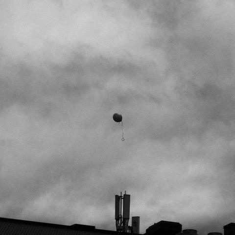 Black and white photo of a lone balloon floating above a building under a cloudy sky.
