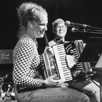 A black and white photo of two people performing on stage. In the front left, a woman with long hair plays a banjo and laughs. In the center, slightly out of focus, a person with short hair and flannel laughs while they play accordion and look on.