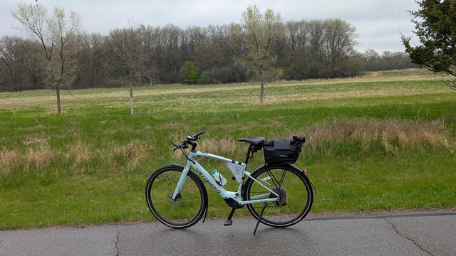 Sage green Vado SL ebike parked on the Bear Creek Trail near the dog park looking east across an open marshy field with greening grass, scattered trees just starting to leaf out and a stand of mostly bare trees in the distance under cloudy skies.