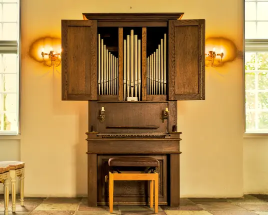 Zwischen zwei hohen Fenstern steht eine kleine Orgel. Die Türen der Orgel sind geöffnet und man sieht die Orgelpfeifen. Vor der Orgel steht ein Hocker für den Organisten. Links und rechts der Orgel hängen Leuchten an der Wand, die das Bild in goldenes Licht tauchen. Links im Raum sind noch zwei Stühle zu erkennen. 
Die Orgel steht in der Kappelle des Schlosses vor Husum. 
. 
A small organ stands between two high windows. The organ doors are open and one can see the organ pipes. There is a stool for the organist in front of the organ. Lights hang on the wall to the left and right of the organ, bathing the picture in golden light. Two chairs can just be recognised on the left of the room. 
The organ can be seen in the chapel of the castle outside Husum. 
.