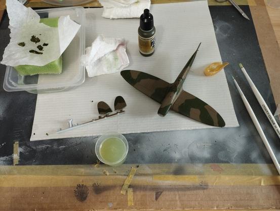 Maqueta a escala 1/72 de un Spitfire MkI. Se le acaba de aplicar una seguna mano de verde oscuro. Enbla imagem se ve la maqueta, un par de pinceles a la derecha, el bote de pintura Vallejo y un bote con agua acompañando a la maqueta en el centro y a la derecha una paleta húmeda casera.