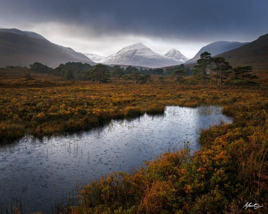 Raindrops cover the surface of a pond surrounded by autumnal heather and grasses. The scene is framed by a triangular wintery mountain peak basking in morning light and a scattering of ancient pine trees.