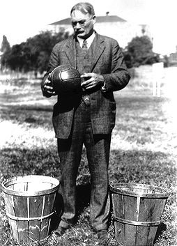 A vintage black and white photograph shows James Naismith, the inventor of basketball, standing between two wooden peach baskets. He is dressed in a suit and tie, holding a basketball. The image, courtesy of Library and Archives Canada (C-80002), depicts Naismith during his time as an instructor at the YMCA International Training School in Springfield, Massachusetts.